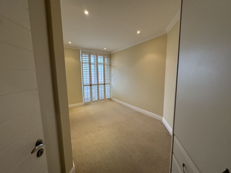 A bright, empty bedroom featuring neutral carpeting, built-in storage cupboards, and white plantation shutters, offering a clean and versatile living space.