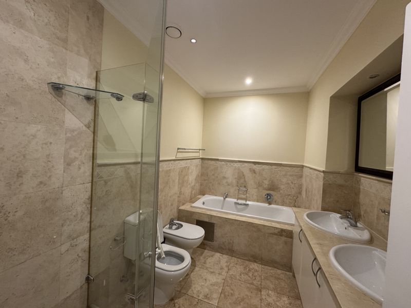 Spacious bathroom featuring a double vanity, built-in bathtub, glass-enclosed shower, bidet, and toilet, finished with elegant travertine tiling throughout.
