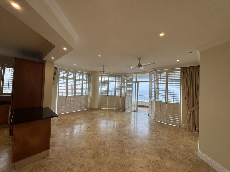 Spacious open-plan living area featuring tiled floors, recessed lighting, ceiling fans, and plantation shutters with direct access to a balcony and ocean views.
