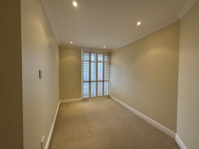 An empty bedroom featuring neutral walls, carpet flooring, recessed downlights, and plantation shutters providing natural light control.