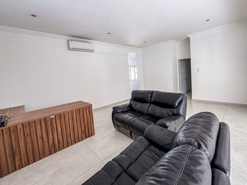 A spacious living room featuring neutral tiled flooring, coved cornicing, recessed downlighting, and a wall-mounted split-system air conditioning unit.