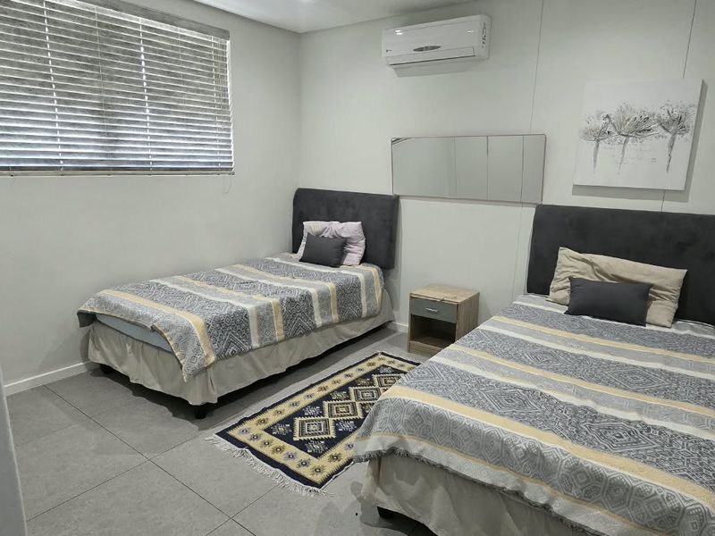A clean, functional bedroom featuring tiled flooring, a wall-mounted air conditioning unit, and a window with venetian blinds.