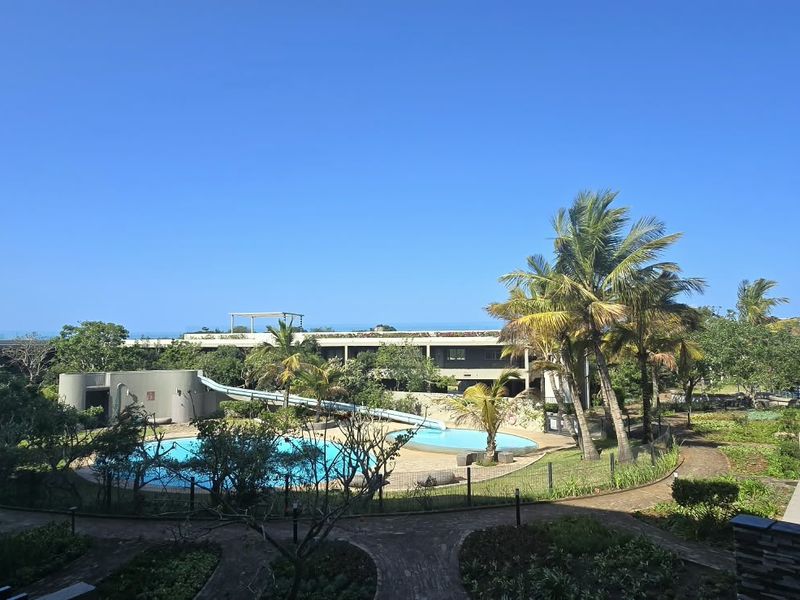 A view of the apartment complex pool area featuring a water slide, landscaped gardens, and paved pathways, viewed from an elevated balcony.