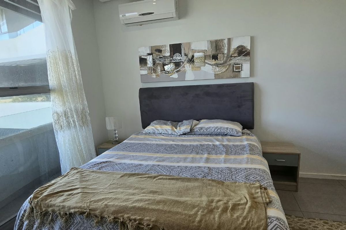 A well-lit bedroom featuring tiled flooring, a large window providing natural light, and a wall-mounted air conditioning unit.
