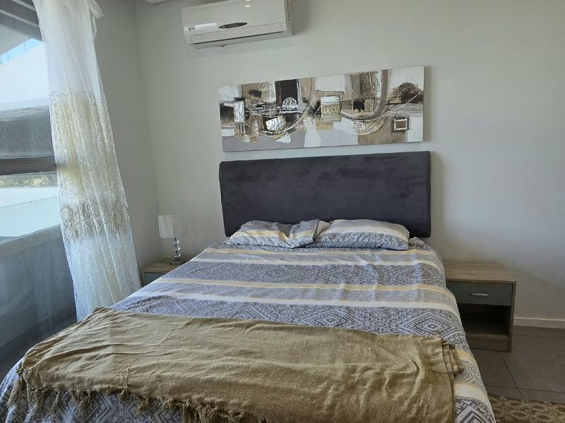 A well-lit bedroom featuring tiled flooring, a large window providing natural light, and a wall-mounted air conditioning unit.