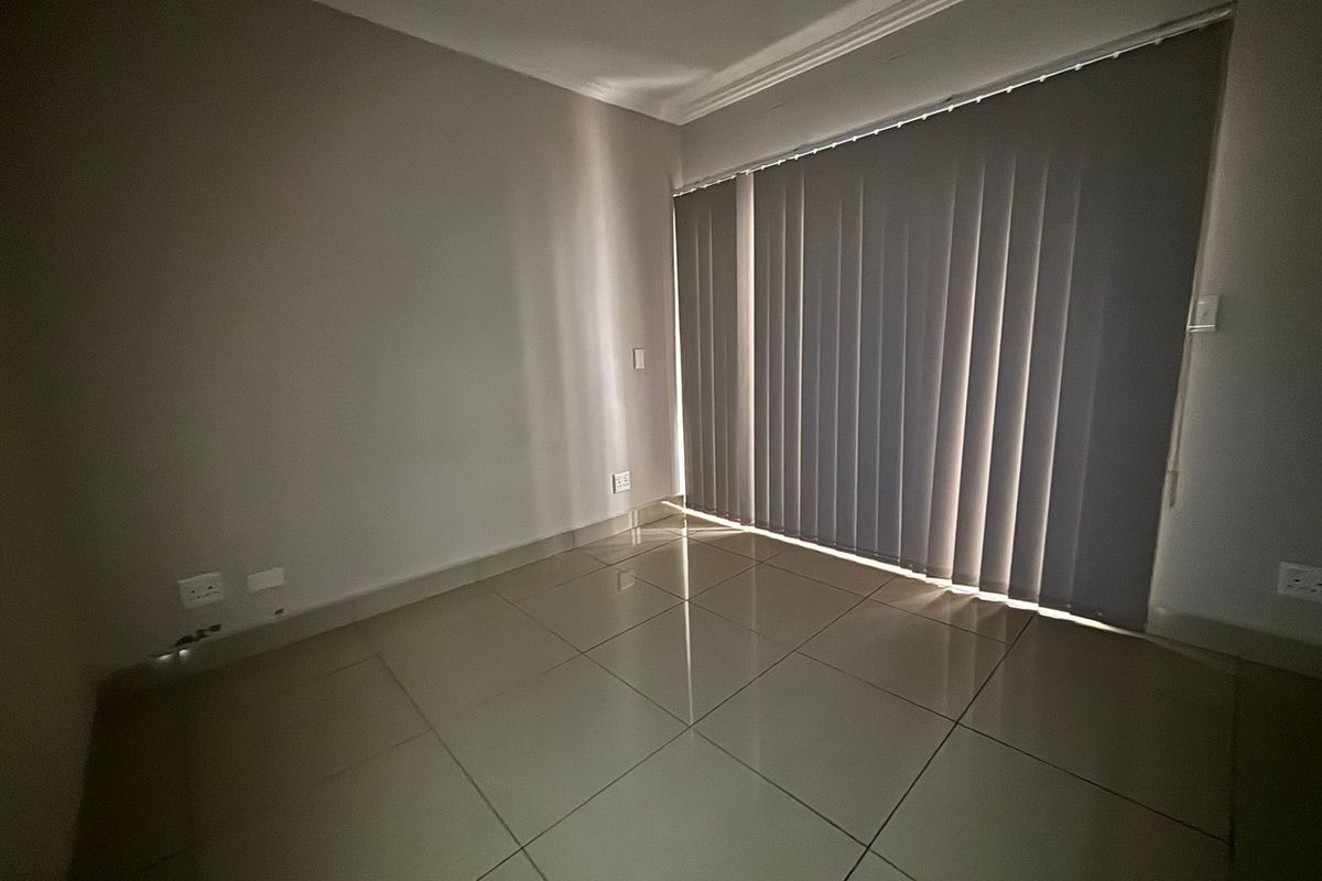 A bright, empty living area featuring neutral tiled flooring, cove cornicing, and vertical blinds covering a large window or sliding door.