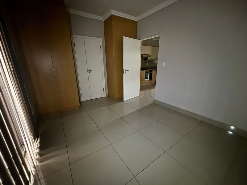 A spacious, empty bedroom featuring neutral tiled flooring, built-in wooden cupboards, and direct access to the kitchen area.
