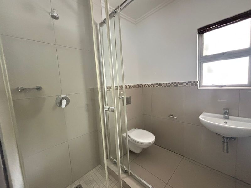 A clean, modern bathroom featuring a glass-enclosed shower, wall-mounted toilet, and a wall-mounted basin with neutral floor-to-ceiling tiling.