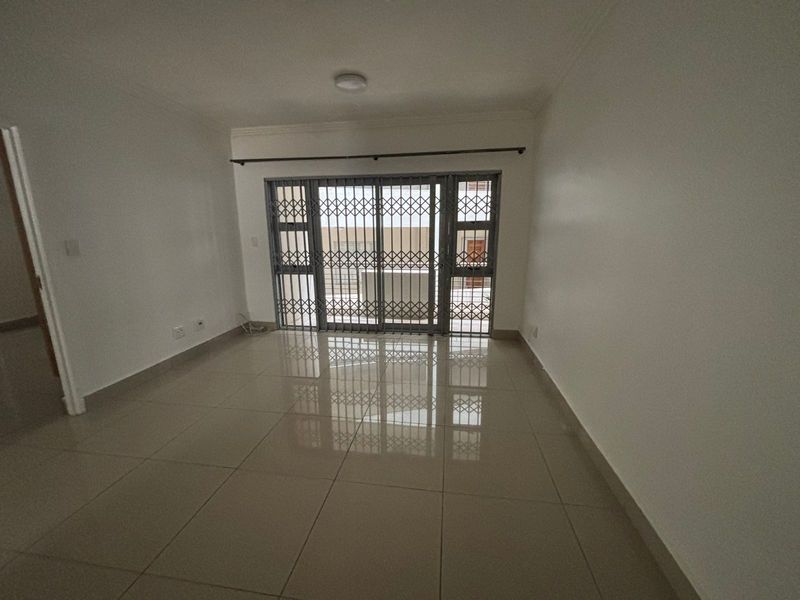 A bright, empty living room featuring neutral porcelain floor tiles, a sliding glass door with a security gate, and clean white walls.