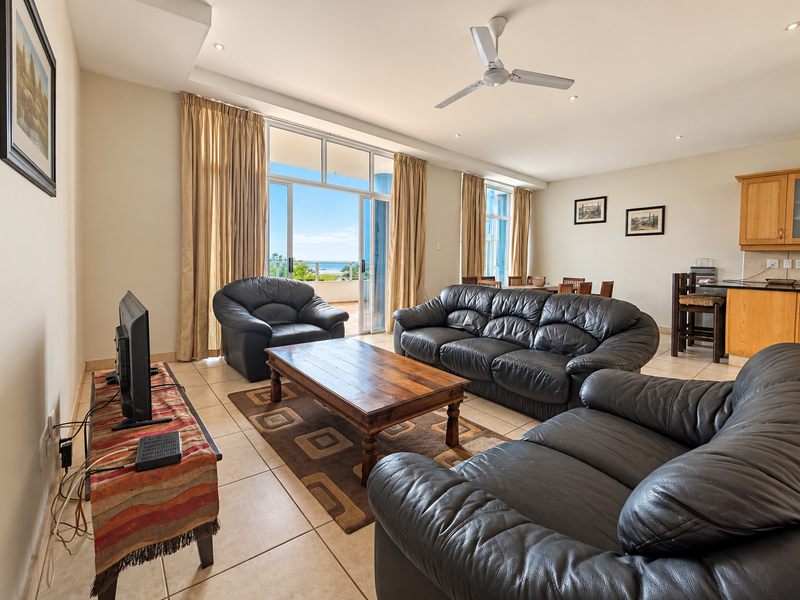 Spacious open-plan living and dining area featuring tiled floors, a ceiling fan, and large sliding doors opening to a balcony with ocean views.