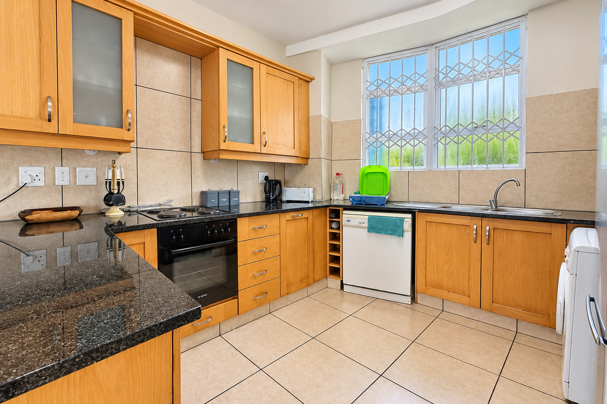 A functional kitchen featuring wooden cabinetry, granite countertops, built-in oven, electric hob, and tiled finishes with a window secured by metal bars.