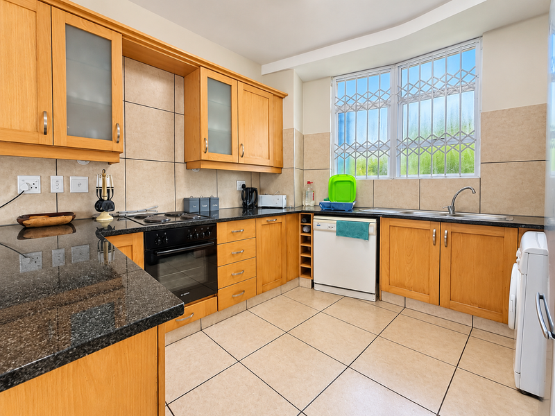 A functional kitchen featuring wooden cabinetry, granite countertops, built-in oven, electric hob, and tiled finishes with a window secured by metal bars.