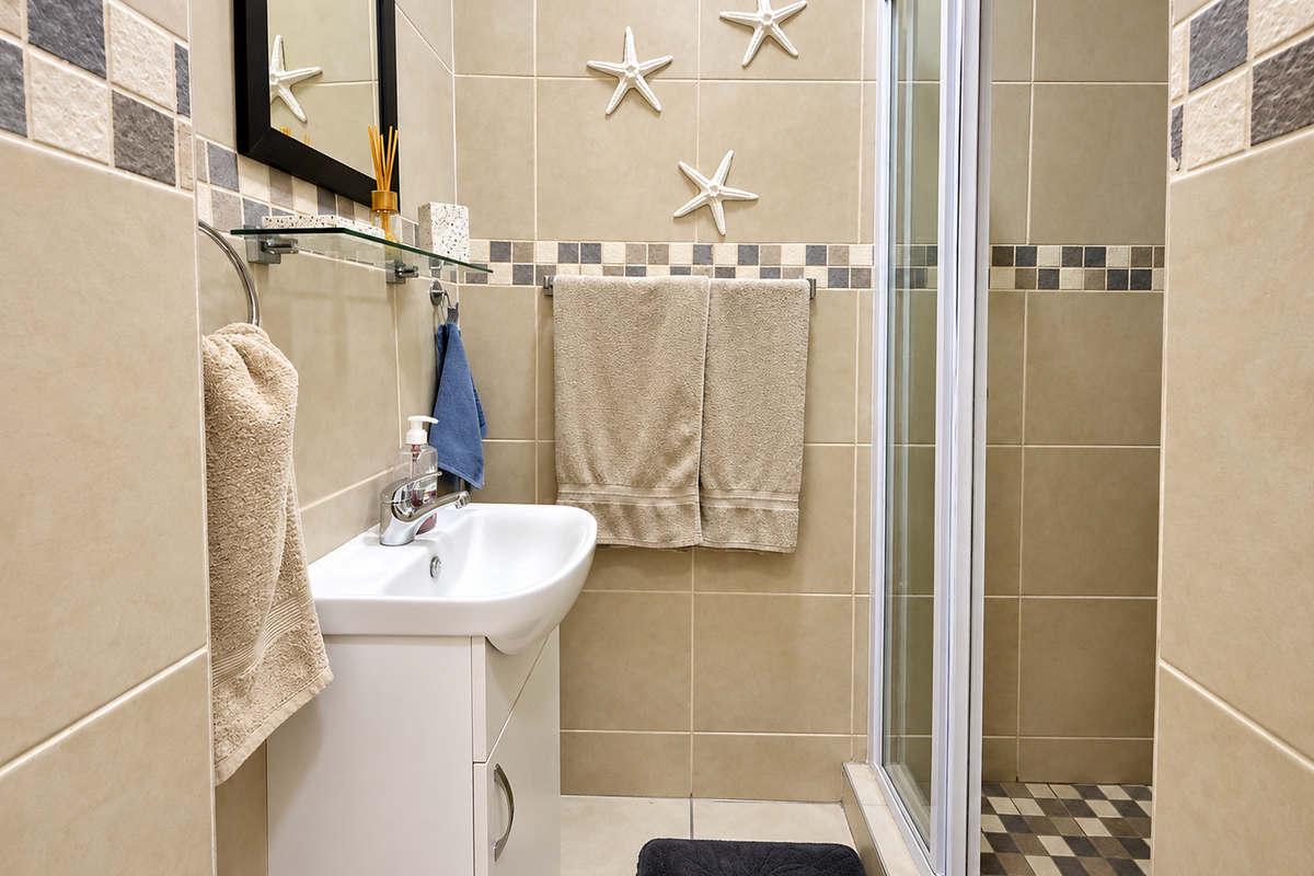 A compact bathroom featuring a vanity basin, a glass-enclosed shower, and neutral-toned wall and floor tiling with decorative accents.