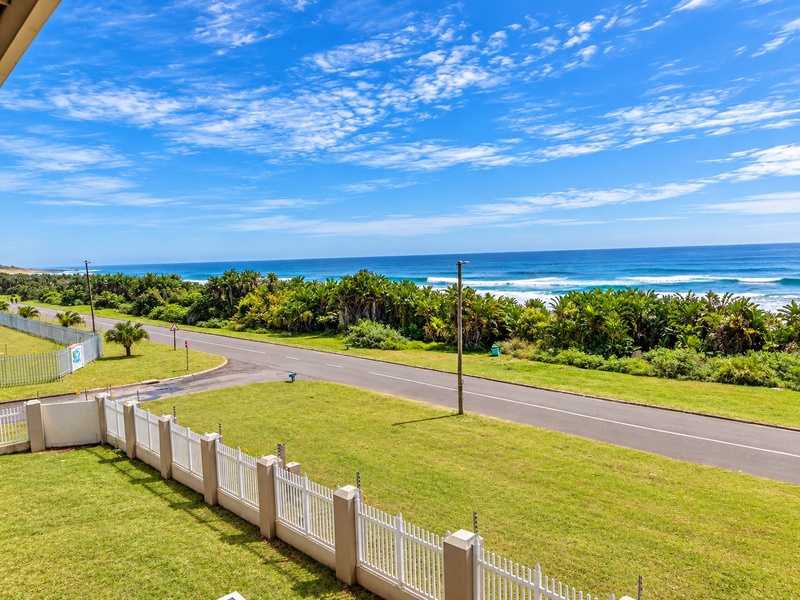Elevated coastal view overlooking a landscaped lawn, perimeter fencing, a quiet road, and the ocean horizon.