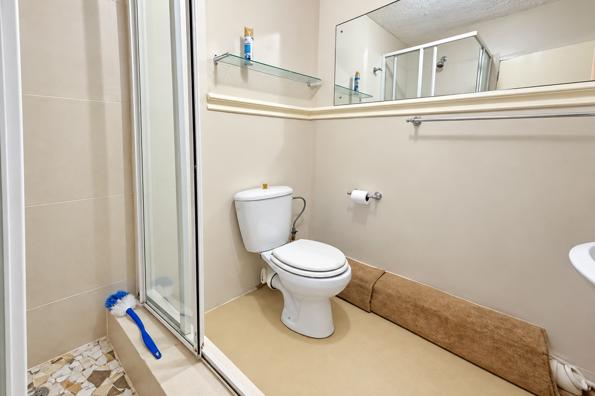 A functional bathroom featuring a shower enclosure, toilet, wall-mounted mirror, and glass shelving, requiring cosmetic updates to the flooring and finishes.