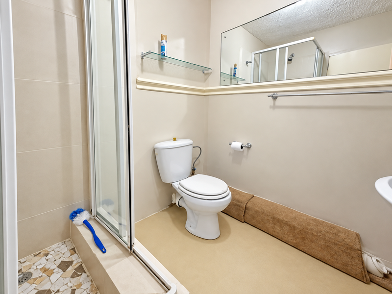 A functional bathroom featuring a shower enclosure, toilet, wall-mounted mirror, and glass shelving, requiring cosmetic updates to the flooring and finishes.