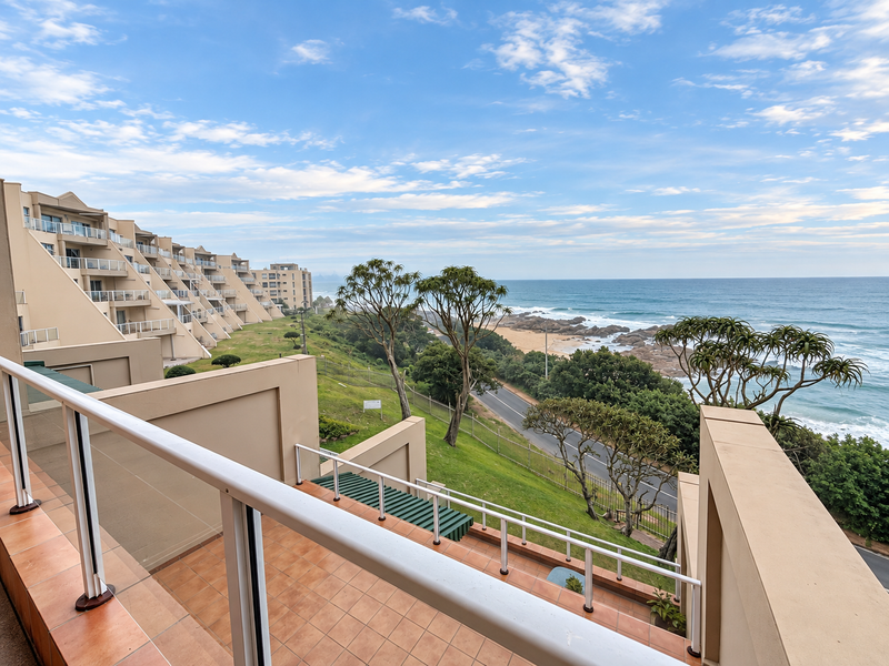 A scenic elevated view overlooking the coastline, beach, and ocean, captured from a private apartment balcony with tiled flooring and safety railings.
