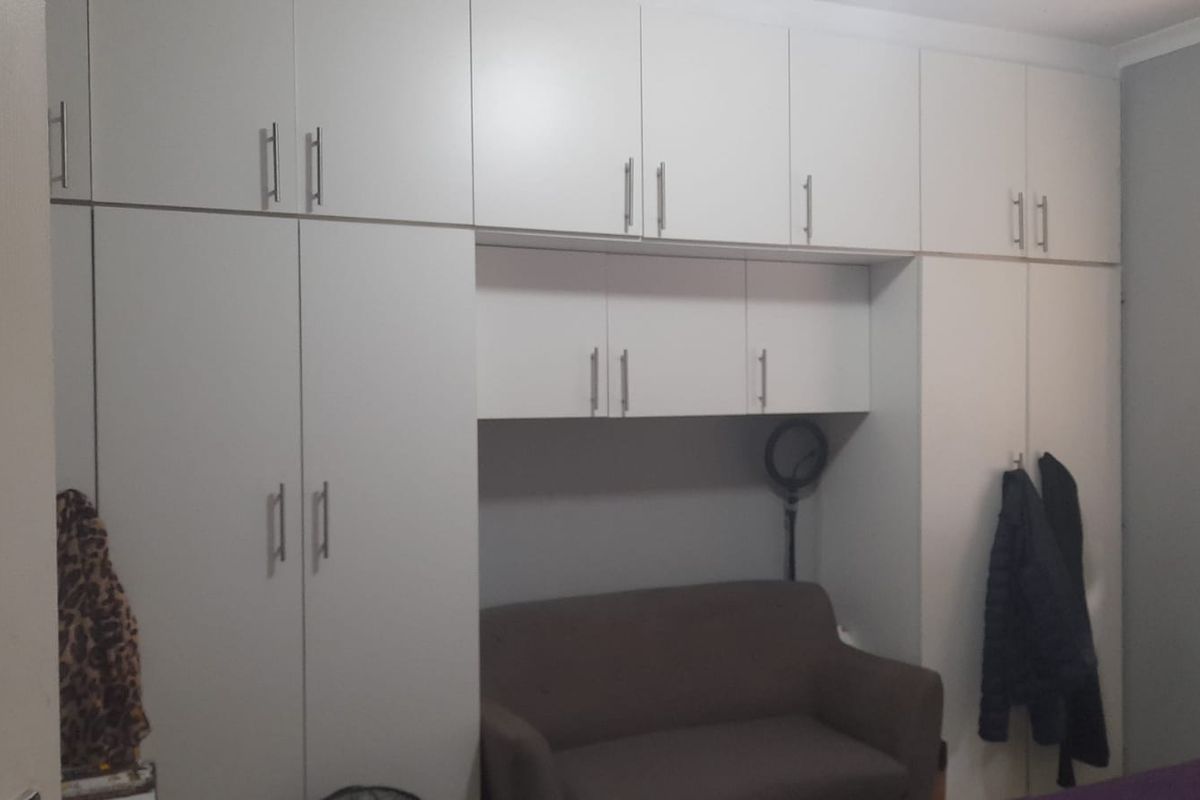A bedroom featuring extensive white built-in cupboards with overhead storage units and a window fitted with venetian blinds.