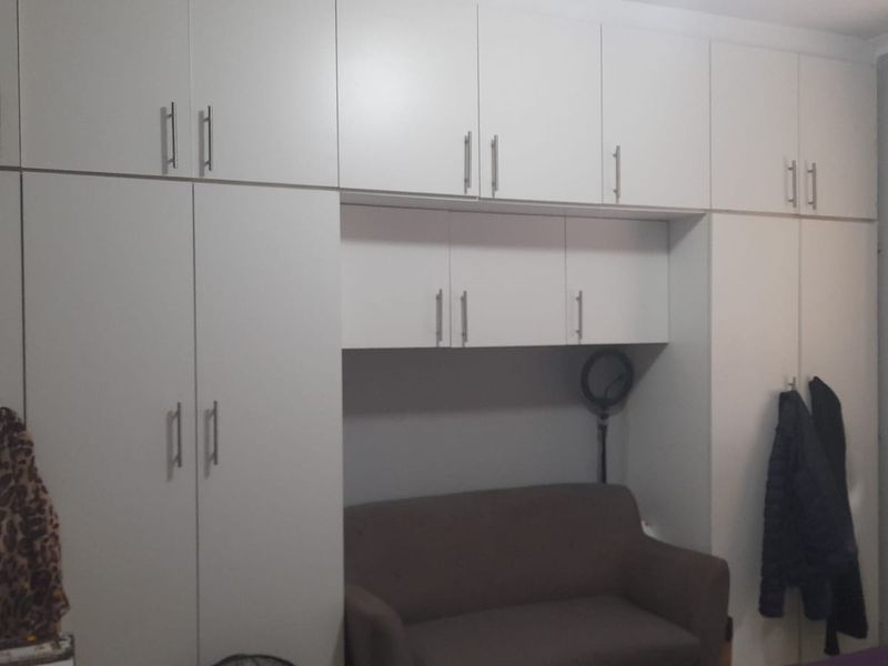 A bedroom featuring extensive white built-in cupboards with overhead storage units and a window fitted with venetian blinds.