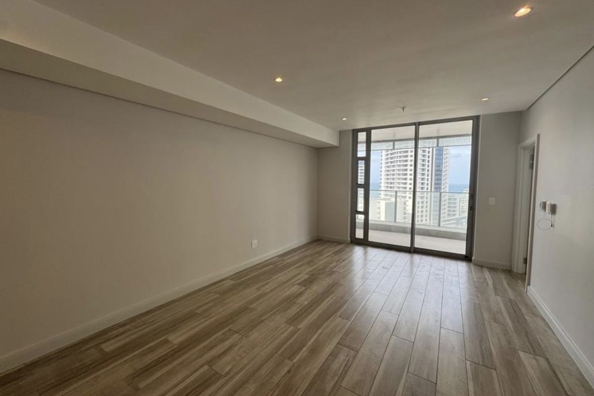 A bright, empty living room featuring modern wood-look tile flooring, recessed lighting, and a large sliding glass door leading to a balcony with city views.