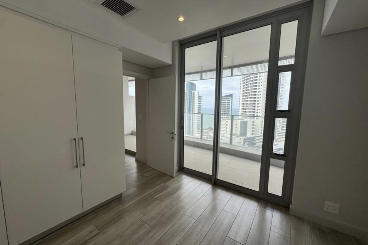 A bright, empty bedroom featuring wood-look tile flooring, a built-in wardrobe, and large sliding glass doors opening onto a balcony with city views.