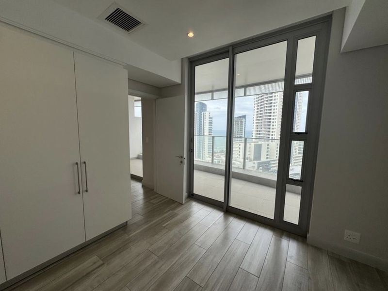 A bright, empty bedroom featuring wood-look tile flooring, a built-in wardrobe, and large sliding glass doors opening onto a balcony with city views.