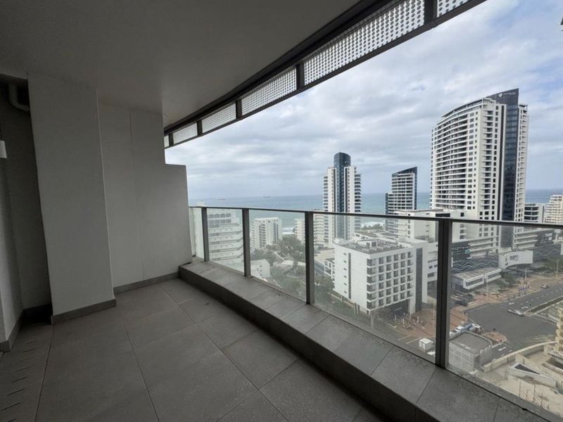 A spacious covered balcony featuring a modern glass balustrade and tiled flooring, offering expansive urban and coastal views.