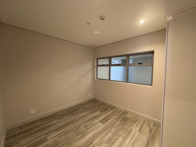 A clean, empty bedroom featuring modern laminate flooring, neutral walls, built-in storage, and an aluminium-framed window providing natural light.