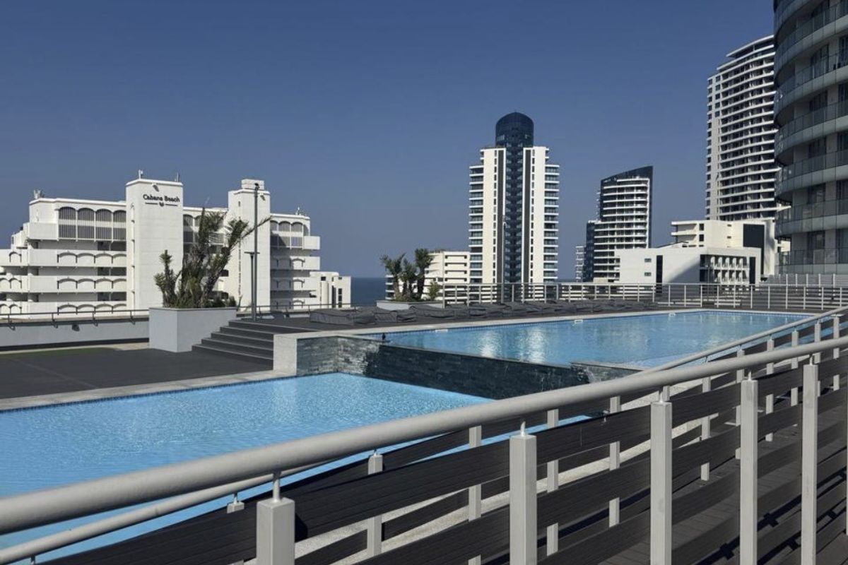 A well-maintained outdoor swimming pool area featuring modern decking, stone accents, and metal safety railings with views of surrounding high-rise buildings.