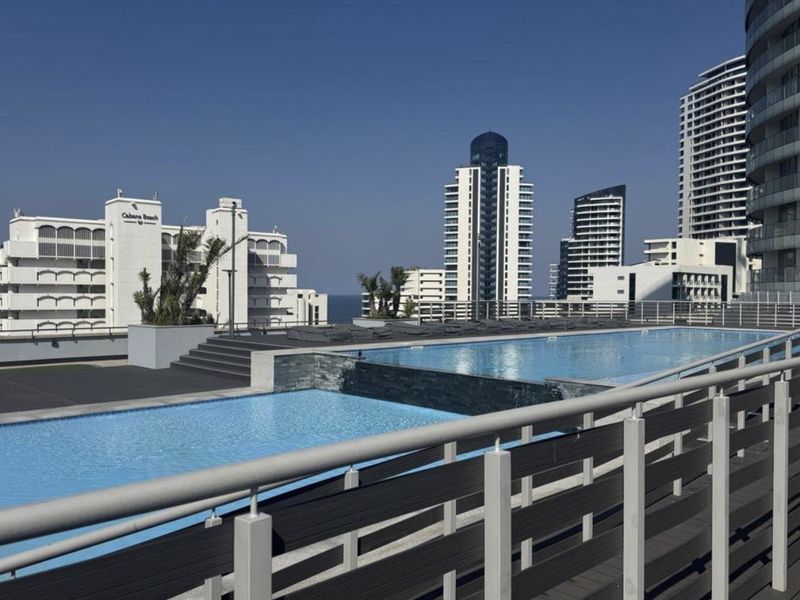 A well-maintained outdoor swimming pool area featuring modern decking, stone accents, and metal safety railings with views of surrounding high-rise buildings.