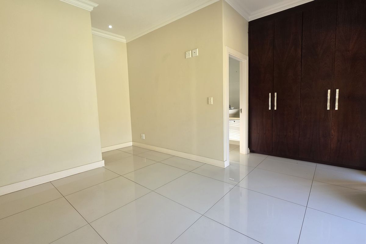 A bright, empty bedroom featuring large format floor tiles, neutral wall finishes, and dark wood built-in cupboards with modern hardware.
