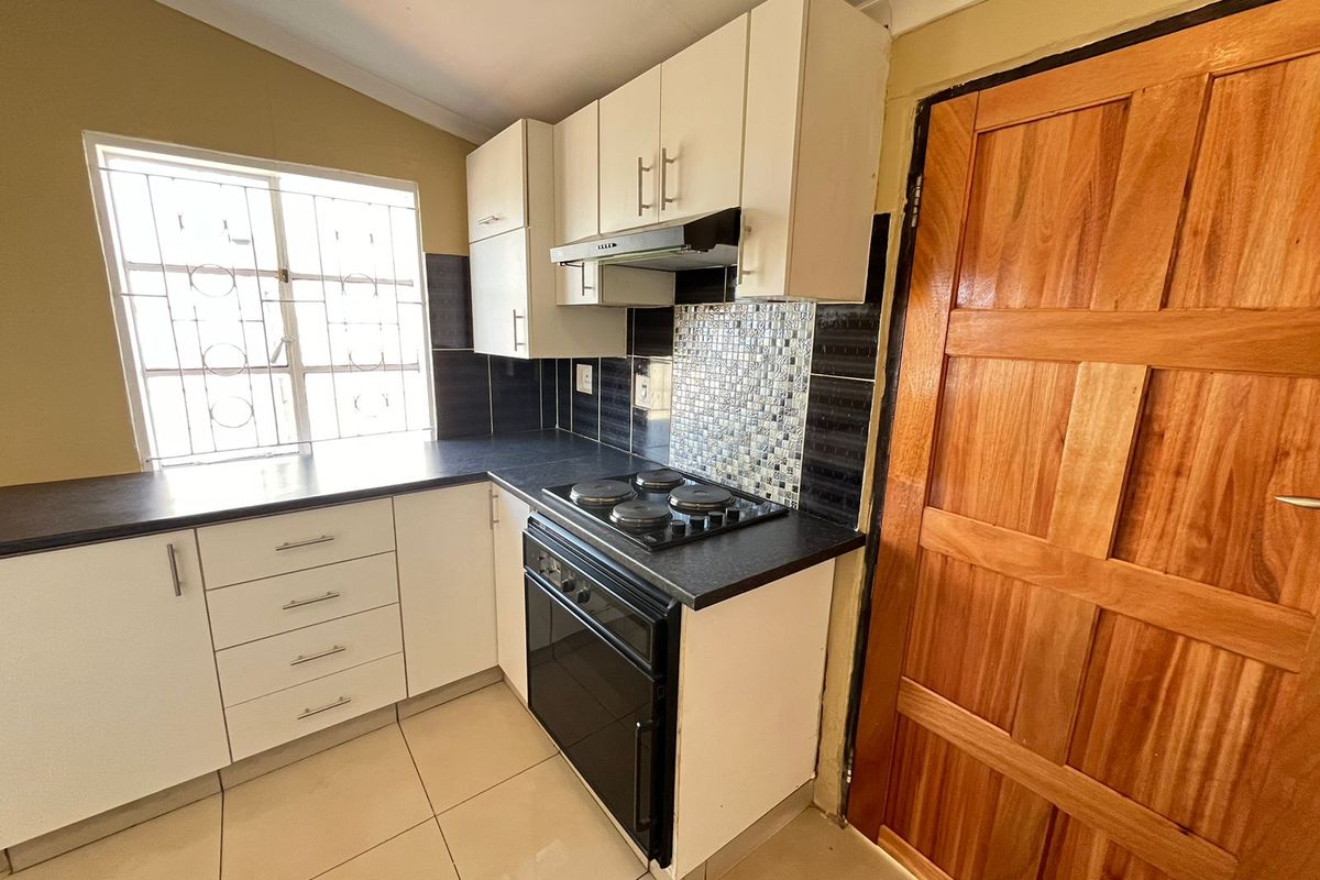 Compact kitchen featuring white cabinetry, black laminate countertops, built-in oven with hob, extractor fan, and tiled splashback with security bars on the window.