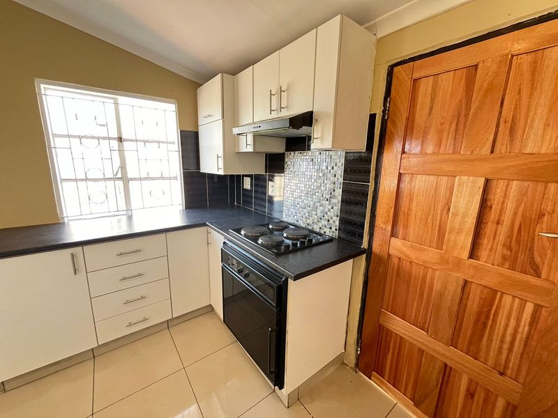 Compact kitchen featuring white cabinetry, black laminate countertops, built-in oven with hob, extractor fan, and tiled splashback with security bars on the window.