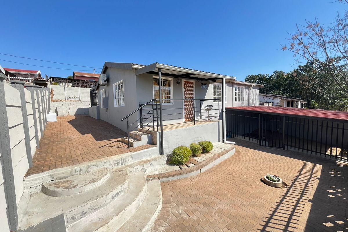 Rear exterior view of the house featuring a paved courtyard, covered patio area, concrete steps, and secure perimeter fencing.