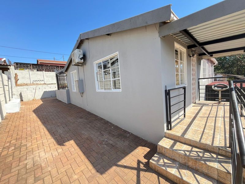 Rear exterior view featuring a paved patio area, tiled steps with metal railings, and a covered patio section with security-fitted windows.