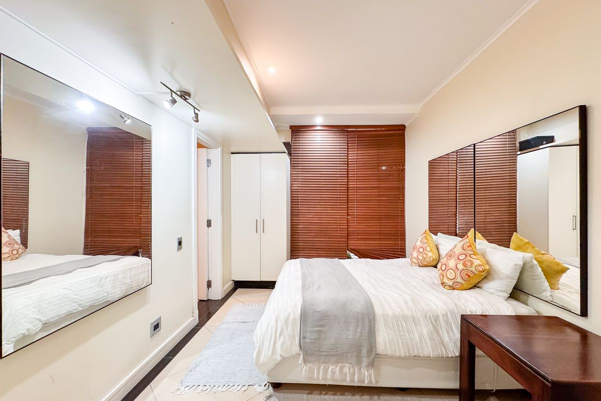 A well-maintained bedroom featuring built-in wardrobes, tiled flooring, and large wall-mounted mirrors, complemented by wooden venetian blinds for privacy.