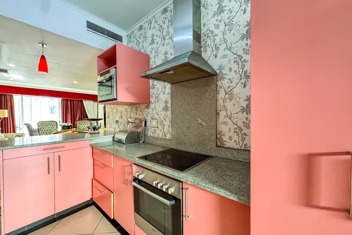 A vibrant kitchen featuring pink cabinetry, granite countertops, a built-in oven, electric cooktop, and stainless steel extractor fan, with patterned wallpaper accents.