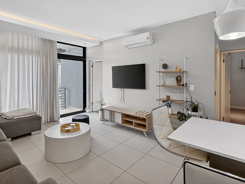 A bright open-plan living area featuring large format floor tiles, recessed ceiling lighting, a wall-mounted air conditioning unit, and access to a private balcony.