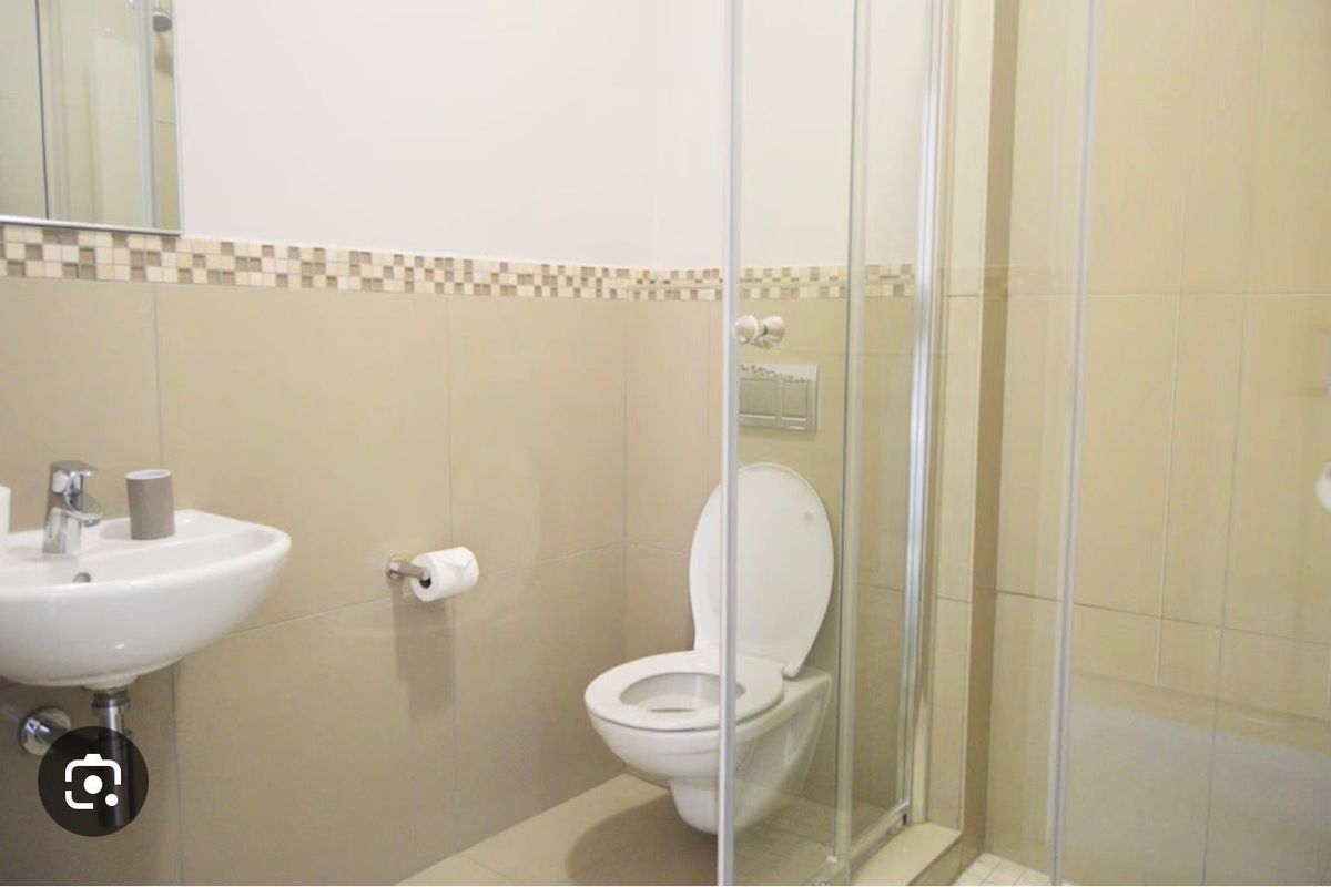 A functional bathroom featuring a wall-mounted toilet, a glass-enclosed shower, and a pedestal basin with neutral wall tiling and decorative mosaic accents.