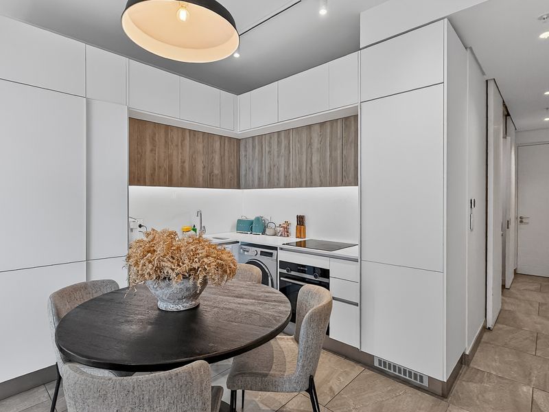 Modern open-plan kitchen featuring sleek white cabinetry, wood-grain accents, integrated oven, cooktop, and built-in laundry facilities, seamlessly connected to the dining area.
