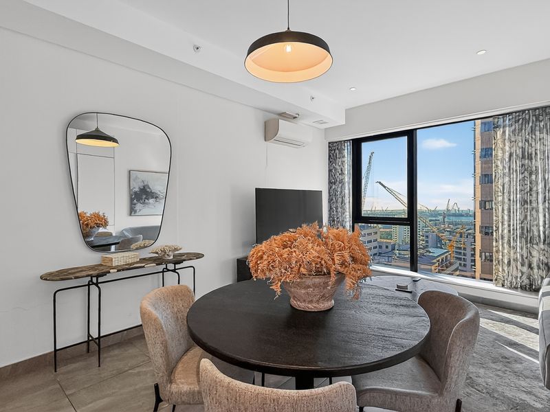 A bright open-plan living and dining area featuring large windows with city views, tiled flooring, and a wall-mounted air conditioning unit.