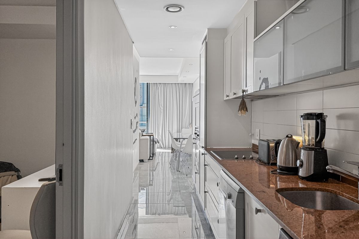 A galley-style kitchen featuring stone countertops, integrated cabinetry, and a stainless steel sink, leading into an open-plan living and dining area.