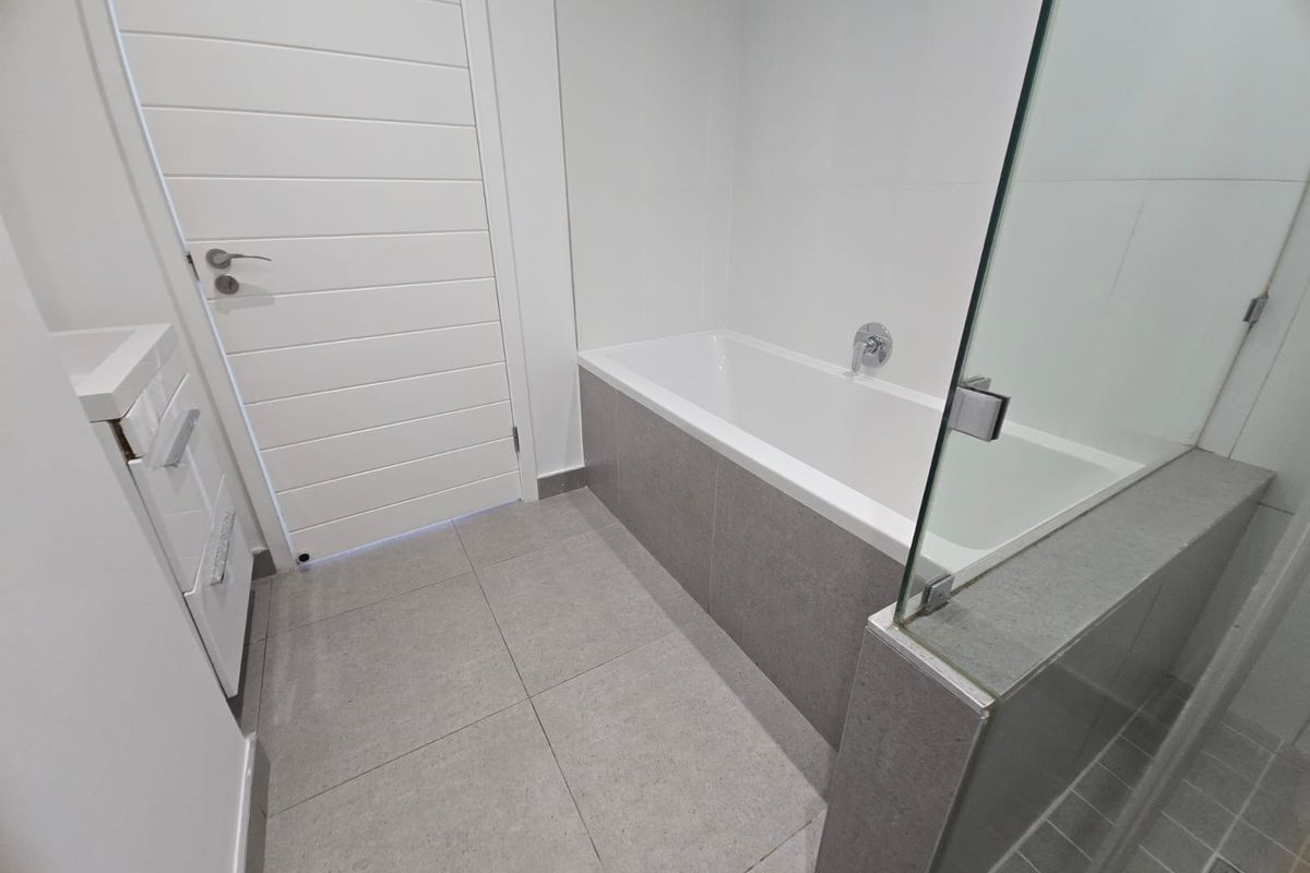 Modern bathroom featuring a built-in bathtub with grey tiled surround, glass shower screen, and tiled flooring.