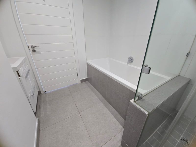Modern bathroom featuring a built-in bathtub with grey tiled surround, glass shower screen, and tiled flooring.