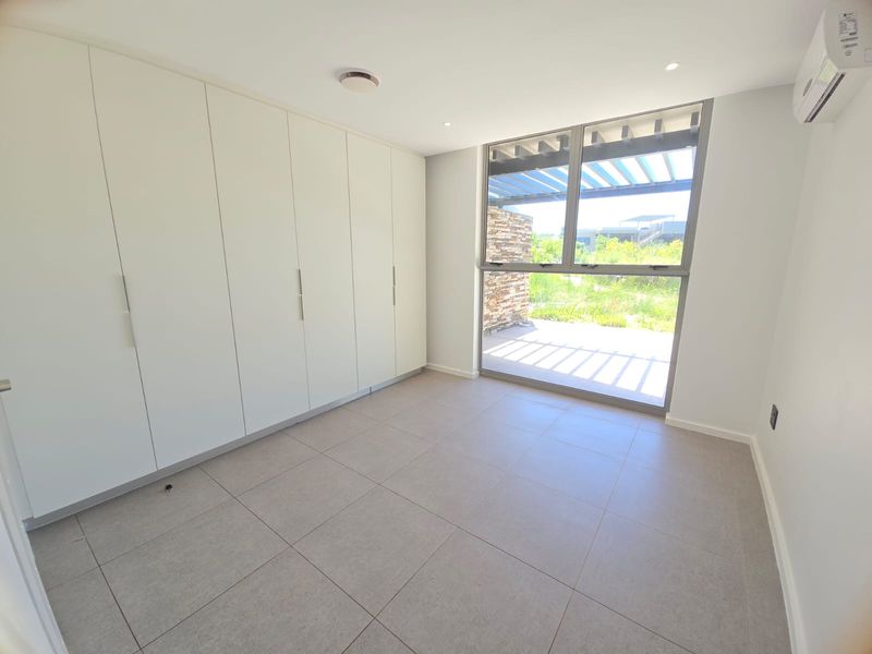A bright, modern bedroom featuring built-in floor-to-ceiling cabinetry, tiled flooring, a wall-mounted air conditioning unit, and large windows with access to an outdoor patio.