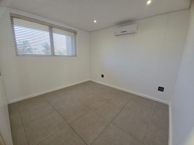 A bright, empty bedroom featuring neutral floor tiles, recessed ceiling lighting, and a wall-mounted air conditioning unit.