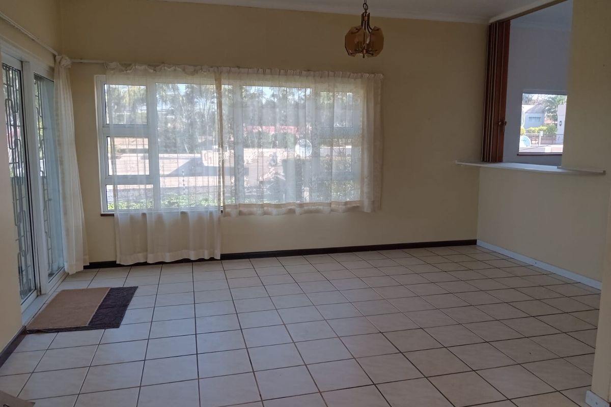 A spacious, empty living area featuring neutral ceramic floor tiles, large windows with natural light, and a pass-through window connecting to the kitchen.