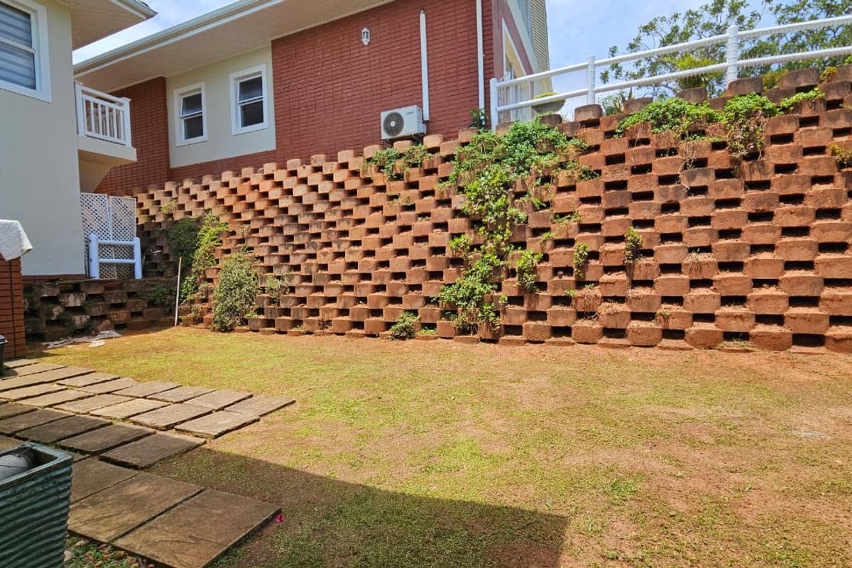 A rear garden area featuring a large tiered concrete block retaining wall, paved walkway sections, and established lawn space.