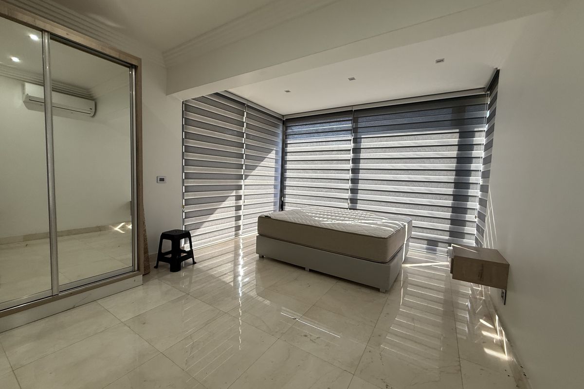 A bright bedroom featuring large windows with modern zebra blinds, polished tile flooring, and a built-in mirrored wardrobe with an integrated air conditioning unit.