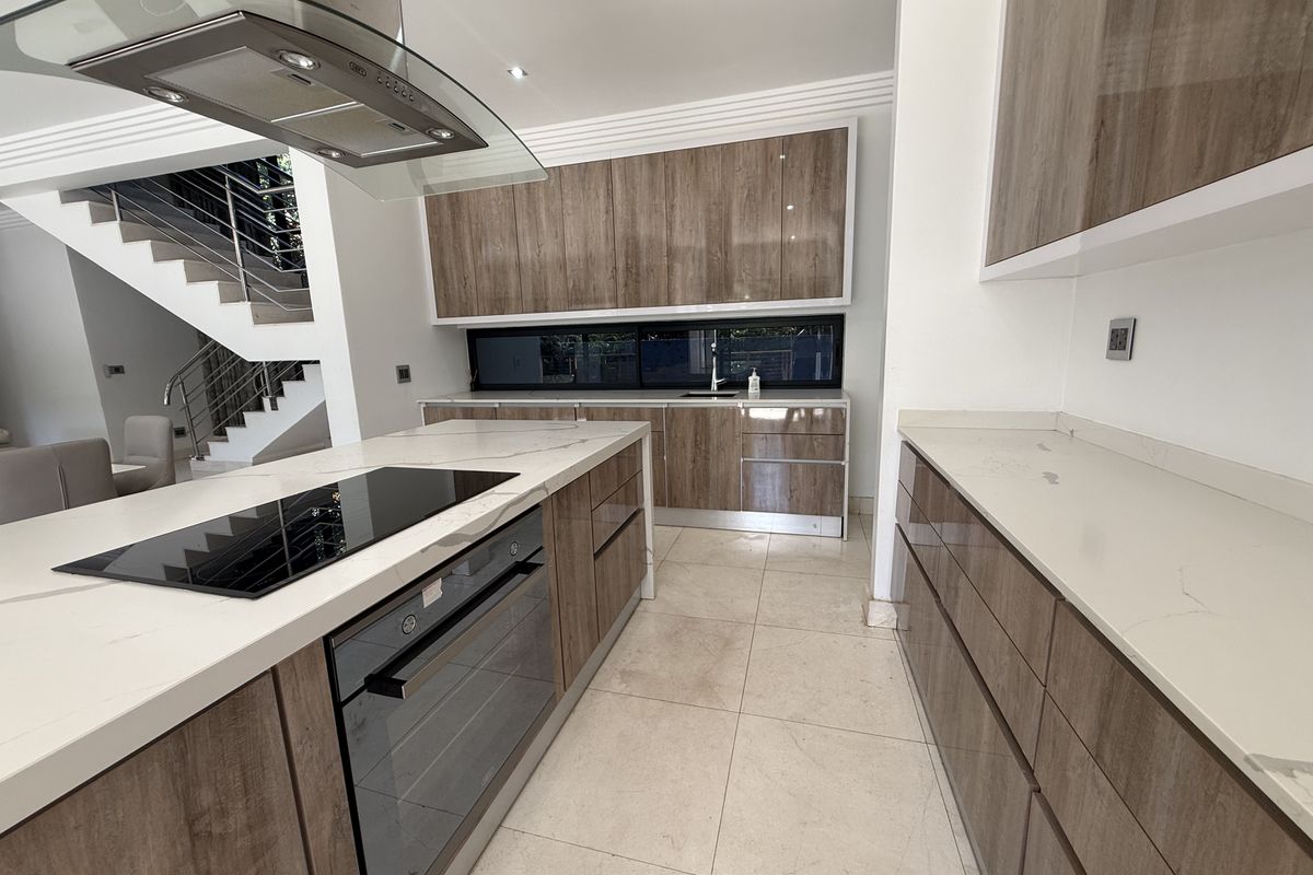 Modern kitchen featuring wood-grain cabinetry, stone countertops, a central island with integrated cooktop and oven, and a sleek glass extractor fan.